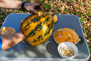 Hands are skillfully carving a pumpkin on a table outdoors, surrounded by autumn leaves, with seeds and pulp collected in bowls nearby.
