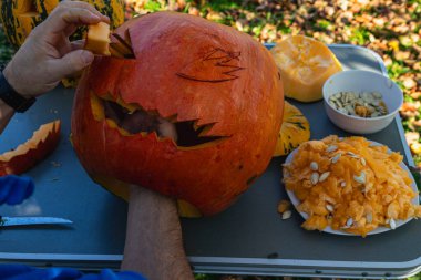 A person is actively carving a pumpkin with a knife in a backyard filled with fallen leaves. Nearby, pumpkin seeds and pulp are neatly arranged on a plate.