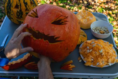 Hands work carefully to carve a jack-o'-lantern with a spooky expression while surrounded by autumn leaves and remnants of pumpkin.