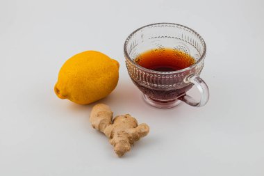 Koyu renkli zencefil çayıyla dolu berrak bir bardak parlak sarı bir limon ve bir parça taze zencefil kökünün yanına yerleştirilir..