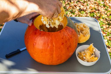 A person is carving a large orange pumpkin outdoors during a sunny day in autumn. Pumpkin seeds and pulp are being placed into a bowl while the background features fallen leaves.