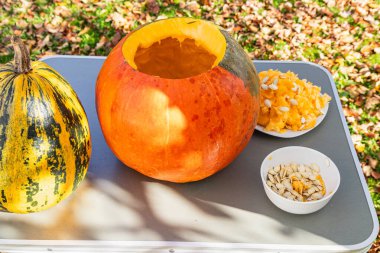 Sonbahar yapraklarıyla dolu canlı bir bahçede balkabakları oyuluyor. İçi boş bir balkabağı, masada sergilenen tohumlar ve hamurla duruyor..