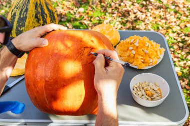 A person sketches designs on a large orange pumpkin while seated at a table covered with colorful gourds and pumpkin guts, enjoying a sunny autumn day.