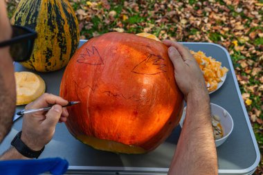 A person sketches a frightening face on a pumpkin in a backyard filled with fallen leaves. Several tools and pumpkin pieces are scattered on the table.