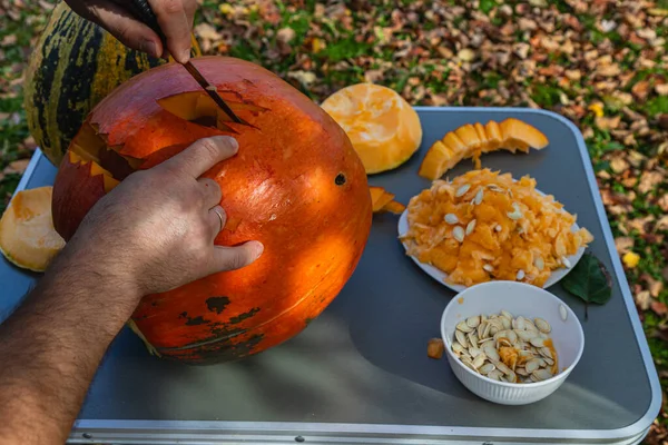 Hands carefully carve a vibrant pumpkin on a table outdoors. Surrounding leaves show the beauty of fall, with pumpkin seeds and scraps adding to the seasonal atmosphere.