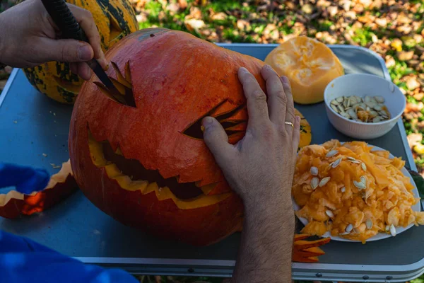 A person carves a jack-o'-lantern outdoors surrounded by fallen leaves and other pumpkins during a sunny fall day, showcasing artistic talent and autumn spirit.