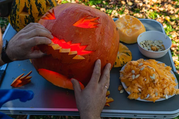 Hands carve a pumpkin into a spooky face, revealing glowing lights inside. Nearby, a plate of pumpkin flesh and seeds sits amongst fallen leaves in autumn.