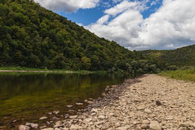 A peaceful riverbank with smooth stones and clear water reflects the blue sky and fluffy clouds. Trees line the hills, creating a calm natural setting for relaxation.