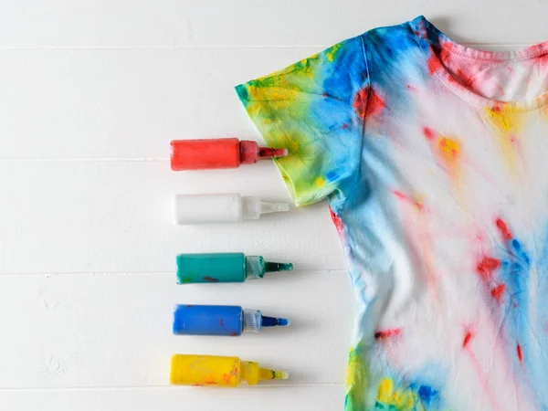 Tubes of paint for clothes and t-shirt in tie dye style on a white wooden table. White clothes painted by hand. Flat lay.