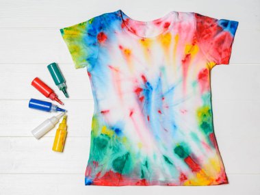 Tubes of paint for clothes and t-shirt in tie dye style on a white wooden table. White clothes painted by hand. Flat lay. clipart