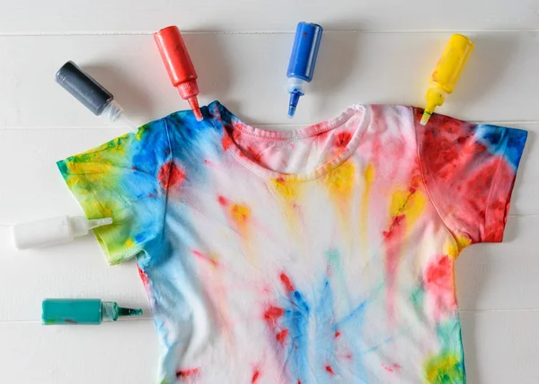 Tubes of paint laid out around the t-shirt in the style of tie dye. White clothes painted by hand. Flat lay.