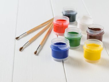 Üç fırçalar ve beyaz ahşap bir masa üzerine guaş yedi kavanoz. Yaratıcı kiti. Hobi ve stil.