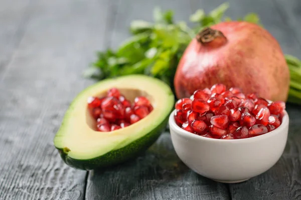 Half avocado with pomegranate seeds, bowl with pomegranate seeds, ripe pomegranate and parsley on a dark table. Ingredients of vegetarian cuisine.
