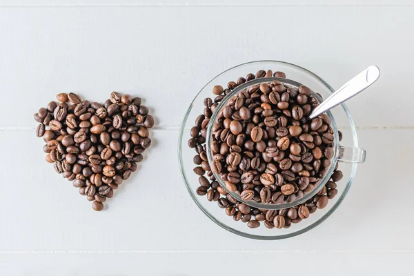 Heart of coffee beans and glass bowl with coffee beans on white wooden table. The view from the top. Flat lay. Grains for the preparation of the popular drink.