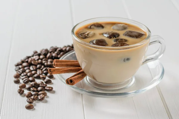 Glass bowl with ice coffee, cinnamon sticks and scattered coffee beans on white table. Refreshing and invigorating drink of coffee beans and milk.