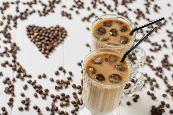 Two glasses of cold coffee on the background of scattered grains in the shape of a heart on a white wooden table. Refreshing and invigorating drink of coffee beans and milk.