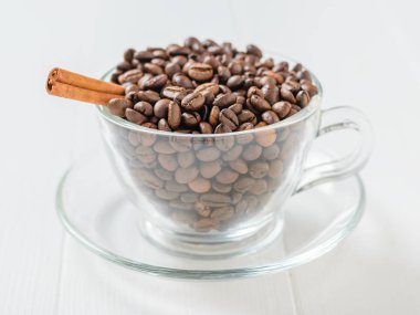 Beyaz ahşap bir masa üzerinde bir cam kabın içinde kavrulmuş kahve çekirdekleri içinde kabuk tarçın. Taneleri popüler içki hazırlanması için.