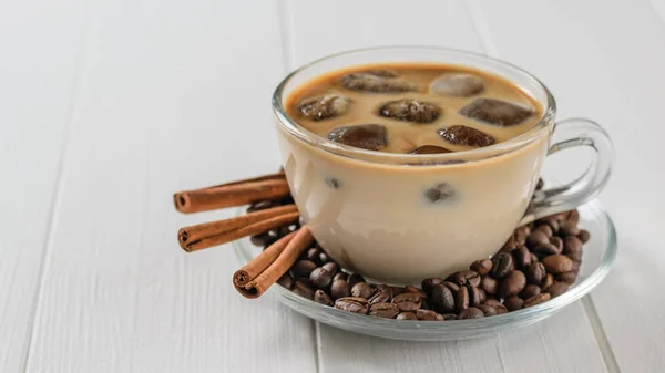 Three cinnamon sticks, a bowl of iced coffee and coffee beans on a white table. Refreshing and invigorating drink of coffee beans and milk.