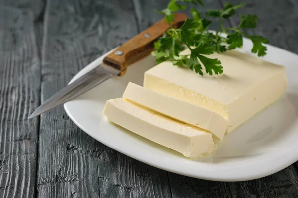 Sliced Serbian cheese with a knife and parsley leaves on a black wooden table. The view from the top. Dairy product.