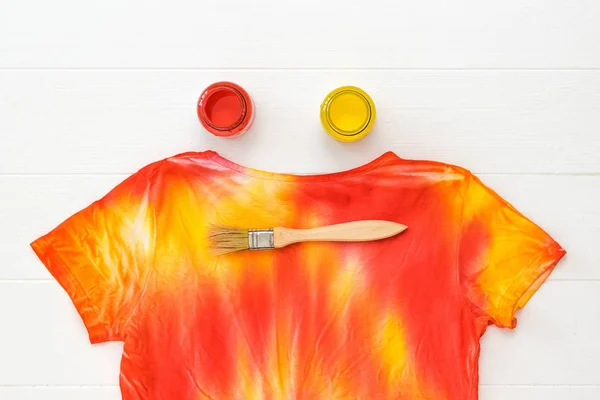 T-shirt painted in the style of tie dye and jars with red and yellow on a white table.