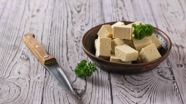 Tofukäsestücke in einer Tonschüssel und ein Messer auf einem Holztisch. Sojakäse. — Stockfoto
