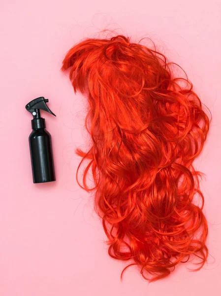 Black hair spray bottle and orange wig on pink background. Accessories to create style.