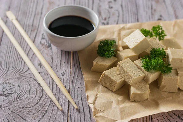 Tofu-Käsestücke auf Papier, eine Schüssel Sojasauce und Holzstäbchen auf dem Tisch. Sojakäse. — Stockfoto