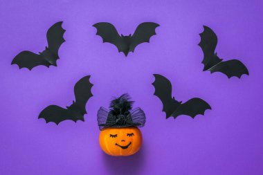 Siyah yarasa siluetleri ve mor arka planda şapkalı bir balkabağı..