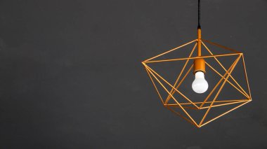 Geometrik metal çerçeveli modern bir kolye ve beyaz bir ampul koyu gri bir tavana asılarak, minimalist bir iç tasarım yaratıyor..