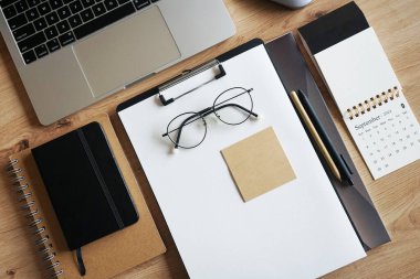 Not defterli ahşap masaüstü, kağıtlı klasör, kağıt, kalem, çıkartma, gözlük, fare ve takvimli dizüstü bilgisayar