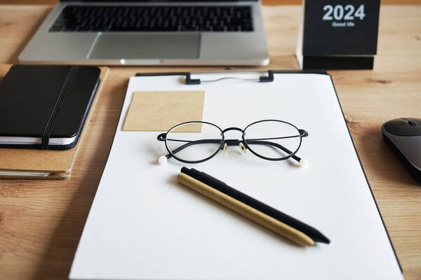 Wooden desktop with notepads, folder with papers, pens, stickers, glasses, laptop with mouse and calendar