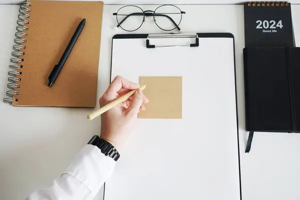 A woman\'s hand holds a pen on the desktop, where there are notepads, paper, stickers, glasses and a calendar