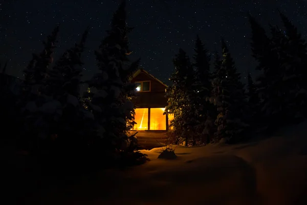 Luz da janela de uma cabana de floresta sob o céu noturno da maneira leitosa na floresta de inverno — Fotografia de Stock