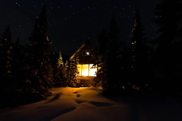 Luz de la ventana de una cabaña forestal bajo el cielo nocturno de la Vía Láctea en el bosque invernal — Foto de Stock