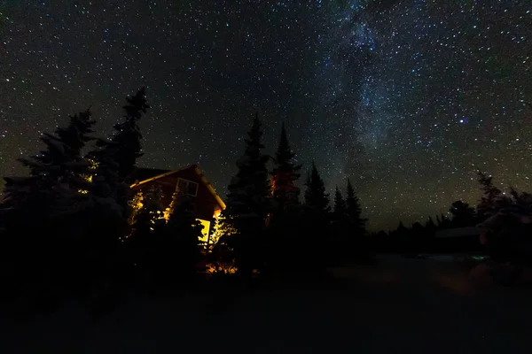 Luz da janela de uma cabana de floresta sob o céu noturno da maneira leitosa na floresta de inverno — Fotografia de Stock