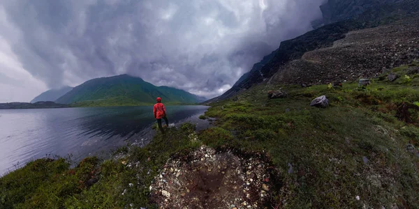 Homme se tient au bord du lac en veste rouge par temps nuageux panorama — Photo