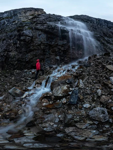 Homem de vermelho fica sob uma cachoeira em uma rocha negra nas montanhas em um dia chuvoso nublado — Fotografia de Stock