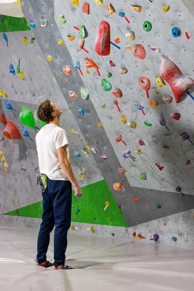 Escalador de roca se para y mira a la pared de escalada con ganchos en la sala de rocas —  Fotos de Stock