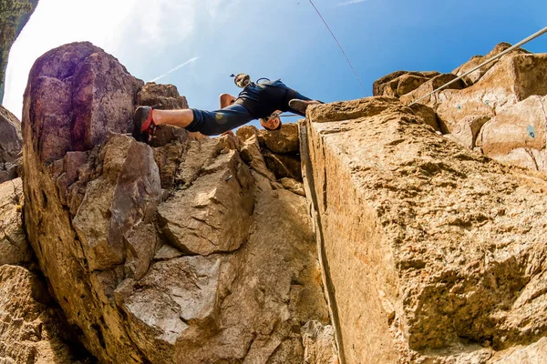 Ακραία ορειβάτης ανεβαίνει ένα βράχο σε ένα σχοινί με την κορυφή, τις ασφαλίσεις το γαλάζιο του ουρανού, κάτω όψη — Φωτογραφία Αρχείου