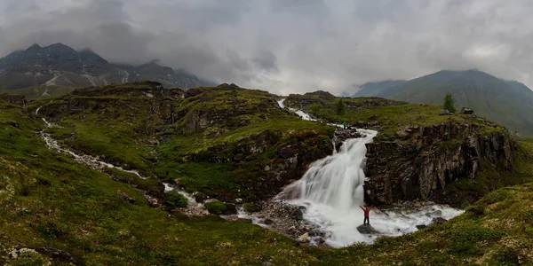 Man in red stands under waterfall on black rock in mountains on cloudy rainy day.. Wide panorama — Stock Photo, Image