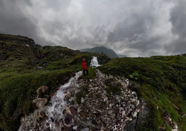 Ο άνθρωπος στο κόκκινο στέκεται κάτω από καταρράκτη σε μαύρο βράχο στα βουνά συννεφιασμένη μέρα βροχερή... Ευρύ πανόραμα — Φωτογραφία Αρχείου