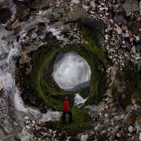 Homme en rouge se tient sous la cascade sur la roche noire dans les montagnes par temps nuageux pluvieux... Petite planète 360 — Photo