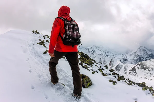 Homem mochileiro no alto em montanhas nevadas. Retrato em um casaco vermelho — Fotografia de Stock