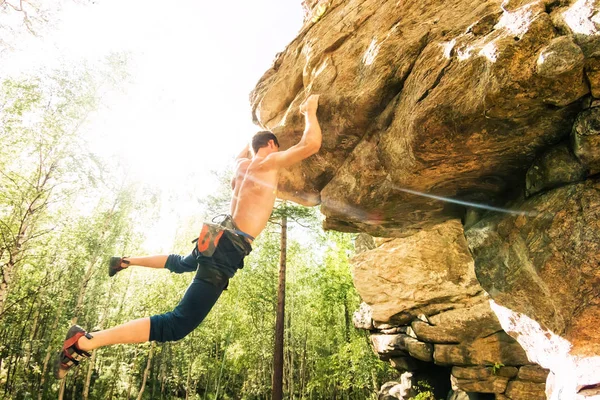 Rock climber climbs bouldering on a cliff on forest. Low angle of strong rock climbing man hanging free on rock with sunflare — Stock Photo, Image