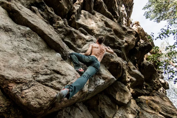 Escalador de rocas sube bouldering en un acantilado en el bosque. Bajo ángulo de fuerte hombre escalador colgando libre en la roca con el sol — Foto de Stock