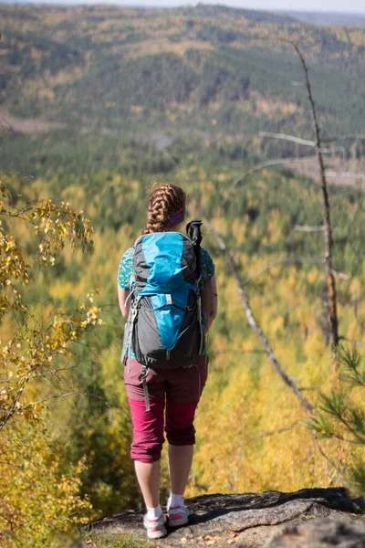 Vörös hajú lány áll egy hátizsákkal egy sziklán az őszi erdő hátterében — Stock Fotó
