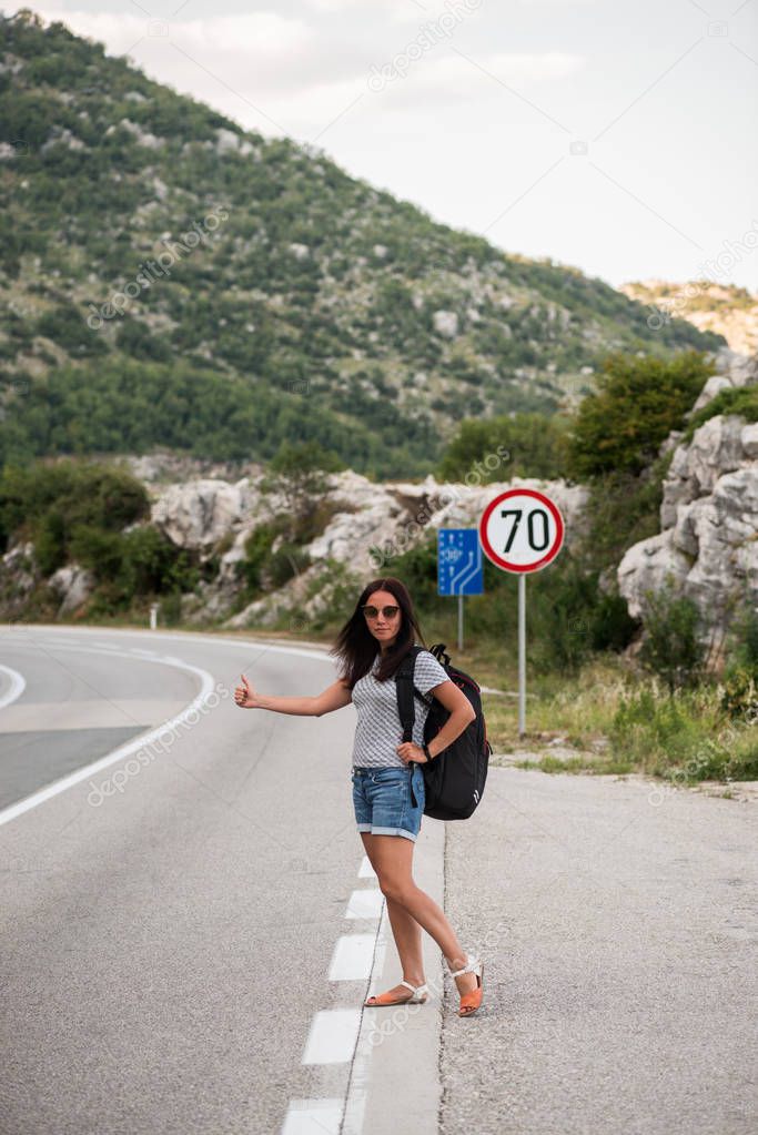 Young beautiful woman with black backpack hitchhiking standing on road. Beautiful young female hitchhiker by the road during vacation trip in mountains.