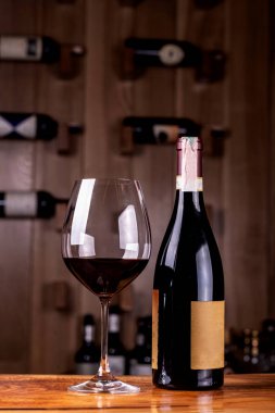 Ahşap masa üzerinde lezzetli kırmızı şarap ile cam ve şişe. Arka planda Kırmızı şarap şişeleri alan sınırlı derinliği ile çekilen ahşap raflar üzerine yığılmış.