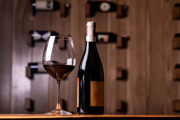 Glas en fles met heerlijke rode wijn op houten tafel. Op de achtergrond rode wijnflessen gestapeld op houten rekken geschoten met beperkte diepte van het veld. — Stockfoto