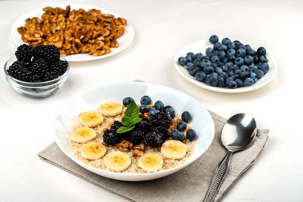 Овсяная каша с бананами, грецкими орехами, ежевикой, черникой, медом и мятой в белой миске на белом фоне. Здоровый завтрак и домашняя диета . — стоковое фото